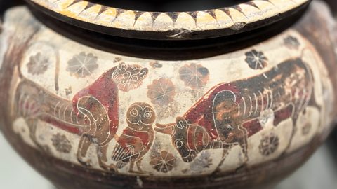 A close‑up of an old painted vase showing animals, including a big bull, a lion‑like creature, and a small owl standing between them. The vase is decorated with flower shapes.