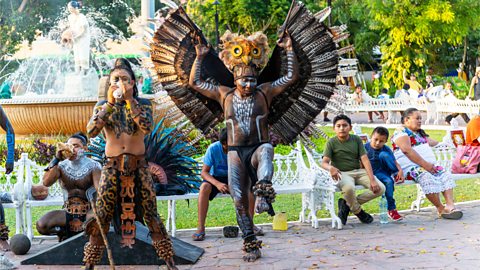 Maya performers in a park are dressed in colorful traditional costumes, including one with large wings.