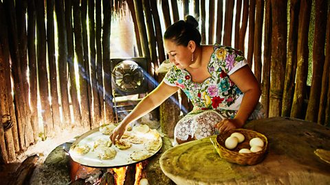 A Maya woman in a colourful dress is cooking round pieces of dough on a big hot pan over a fire inside a small wooden hut. There is a basket of eggs on a nearby table.