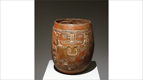 A brown, barrel‑shaped clay pot with carved patterns on the outside. The designs look like simple shapes and faces. The pot is standing on a small white platform in a museum.