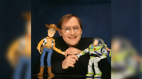 John Lasseter, who has brown hair, glasses and wears a long-sleeved black top holds to hand of two toys, Toy Story characters Woody and Buzz Lightyear. Woody is a cowboy sherriff and Buzz is a space ranger