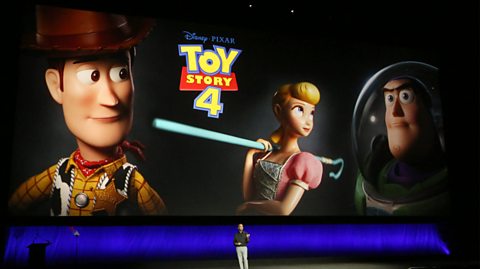 Producer Jonas Rivera talks about the movie Toy Story 4 at CinemaCon in 2019. He is on stage wearing a long-sleeved black top and grey pants. Behind him is a huge screen with the yellow and red Toy Story 4 logo and characters Woody - a cowboy sheriff, Bo Peep - a porcelain shepherdess, and Buzz a space ranger 