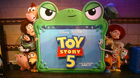 A promotional board for Toy Story 5 shows a number of characters standing around and behind a key character looking worried and scared. The main character is a large green touch-screen table with frog eyes and frog legs called Lilypad