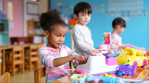 In a classroom with wooden chairs and light blue walls, a little girl with a multicoloured top and curly hair in a bun plays with a plastic cash registered. In the background are two more children, a boy and a girl, playing with other toys on the table