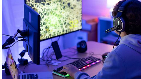 A person is sitting at a multi-screen computer with a light-up keyboard and other technology in front of them. They are wearing a pair of gaming headphones. 