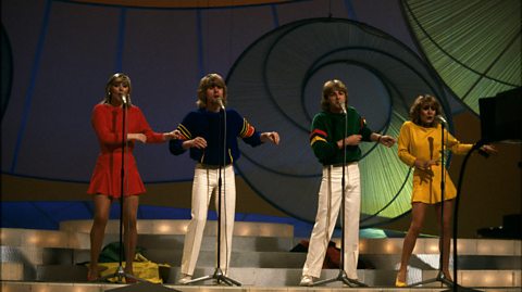 Bucks Fizz perform on stage at Eurovision, dancing behind mics on stands. The girls wear long sleeved tops and skirts, one red and one yellow. The boys in the centre wear white pants and navy jumpers with red and yellow stripes on the arms
