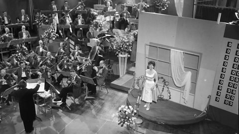 Lys Assia, the first winner of Eurovision, stands on stage in a black and white photo. She wears a white dress with a window display behind her on stage. She is surrounded by an orchestra and flowers