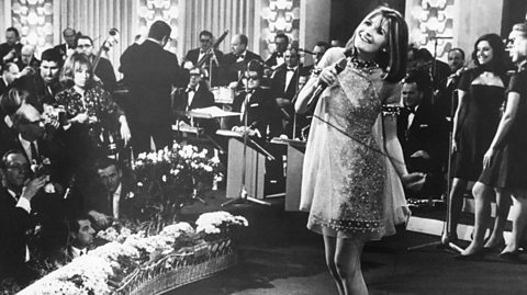 Sandie Shaw, the first UK winner of Eurovision, in a black and white photo. She stands barefoot on stage with a floral shift dress, singing into the mic. Around her is a small orchestra