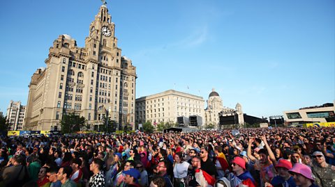 Eurovision fans descend on Liverpool for the final in 2023. The large crowd wearing colourful clothes and flags in the sunshine stand on Liverpool's waterfront, with the Three Graces buildings behind them