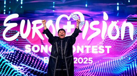 JJ, winner of Eurovision 2025, stands on stage cheering with his arms in the air celebrating. Behind him is the Eurovision Song Contest logo in white with purple and blue lights behind it. He has short dark hair and wears a long sleeved black outfit