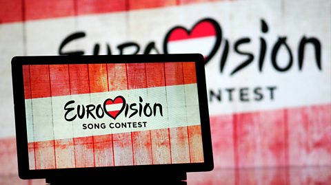 The Eurovision Song Contest logo on a tablet and projected onto a large screen behind it. In 2026, it will take place in Austria. The logo has a heart with the Austrian flag - which is a white stripe in between two red stripes