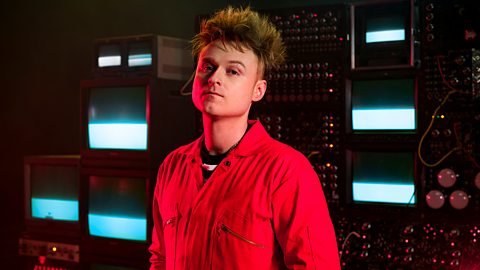 Artist LOOK MUM NO COMPUTER wears a red boiler suit and has spiced blonde hair as he stands in front of technical equipment and televisions on set of his music video
