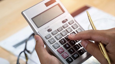 Hands hold a silver calculator and type on the buttons. The right hand also has hold of a gold pen and blurred in the background is paper and a pair of glasses on the table