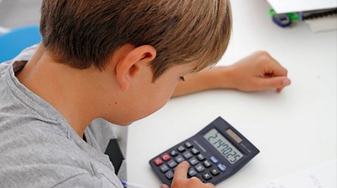 A young boy with blonde hair and a grey top types equations into a navy blue calculator which rests on a white table