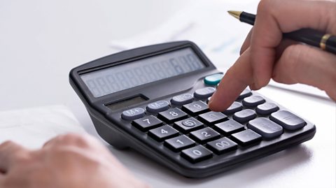 A square grey calculator rests on a white table as a hand holding a pen presses the buttons to make a calculation