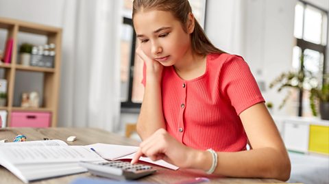 A student using a calculator while studying. She has brown hair and wears a coral short-sleeved buttoned top. On her wooden desk is the grey calculator, and her open study books. 