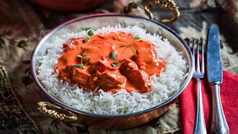An ornate dish filled with rice and chicken tikka masala, with cutlery on a napkin to the side