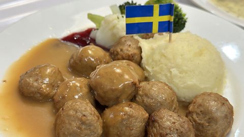 A plate of Swedish meatballs, covered in a gravy, with vegetables and mashed potato. There is a Swedish flag stuck into the potato