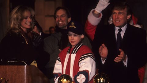 In 1991, Macauley Culkin, wearing a red and white baseball jacket and black cap, stands centrally with Donald Trump stood behind clapping. 