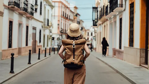 Getty Images Woman wearing a straw hat and backpack walks alone down a quiet street lined with pastel-coloured buildings (Credit: Getty Images