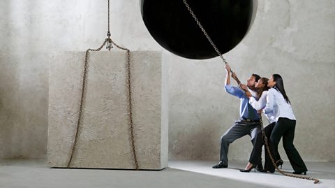 Three office workers in shirts and trousers - two women and one man - hold a chain and look nervous as they are about to pull a huge heavy cube tied to the chain