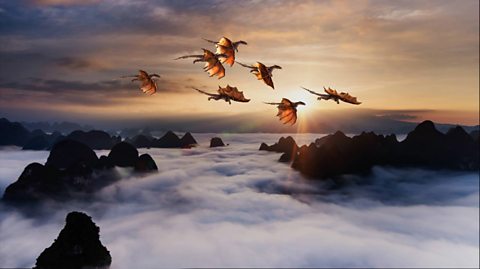A computer generated image of seven orange dragons - large scaled animals with wings - flying over misty mountains