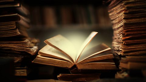 Pages in an open book project light and magic out of them as piles of books rest either side