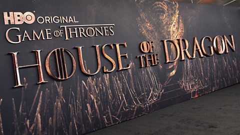 A promotional board for HBO series House of the Dragon. The dark board has an image of a large dragon, a scaly serpent like creature and lots of swords in a large throne room. It has the logo House of the Dragon in bold, brass-like text