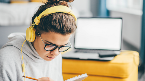 Image of a young woman writing notes on a pad while wearing headphones