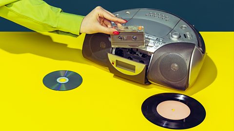 A hand with red painted nails and wearing a green shirt puts a cassette tape into a silver stereo music player. It sits on a yellow table with a CD and a vinyl placed next to it