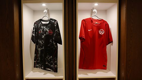 A black and white Canada away shirt, decorated with abstract maple leaf graphics is hung up next to a red home shirt, also with a large maple leaf motif on it