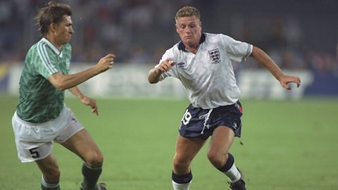 Paul Gascoigne, wearing the white Umbro Italia 90 shirt with navy shorts and white socks, dribbles past a German opponent in a green shirt and white shorts