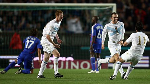 James McFadden celebrates his goal against France with teammates, wearing an all white kit with a sky blue cross over the chest, and gold trim