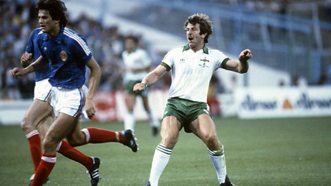Gerry Armstrong of Northern Ireland watches a shot (off camera) in a 1982 World Cup match against Yugoslavia, wearing a white shirt with green pinstripes, green shorts and white socks