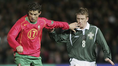 Cristiano Ronaldo, in the red shirt and green shorts of Portugal, wearing 17, dribbles away from Northern Ireland's Steve Davies, wearing 8, in the lace-up Northern Ireland kit
