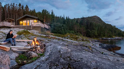 Alamy A woman sits on a remote shore on the Norwegian coastline in front of a campfire, a cabin and a forest while reading a book under a moody grey sky (Credit: Alamy)