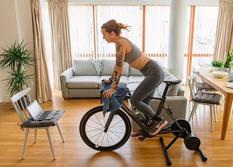 Getty Images A woman pedalling on an exercise bike in her living room with a laptop on a chair in front of her (Credit: Getty Images)
