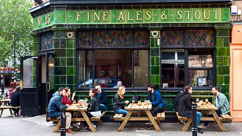 Alamy People sit outside a green-tiled pub in London on wooden benches with gold lettering reading 'Fine Ales and Stout' (Credit: Alamy)