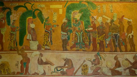 A colourful wall painting shows many people in bright clothes standing, walking, and working. Some carry big green leaves and baskets, and the pictures are arranged in rows like a long story painted on the wall.