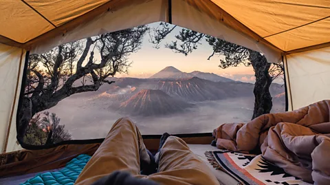 Getty Images Two people lie on sleeping mats in a tent overlooking a mountainous view, with two trees framing the tent canopy in the foreground (Credit: Getty Images)