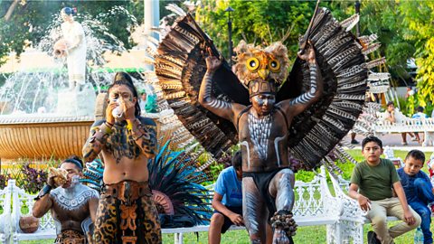 Maya performers in a park are dressed in colourful traditional costumes, including one with large wings.