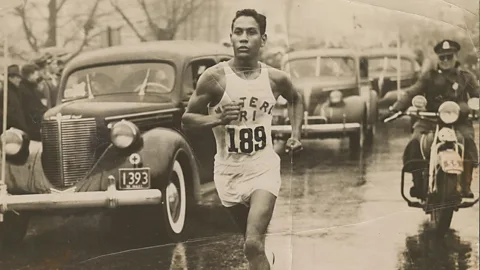 BAA Sepia photograph of Ellison "Tarzan" Brown running down a Boston street with cars and a motorcycle  trailing behind him(Credit: BAA)