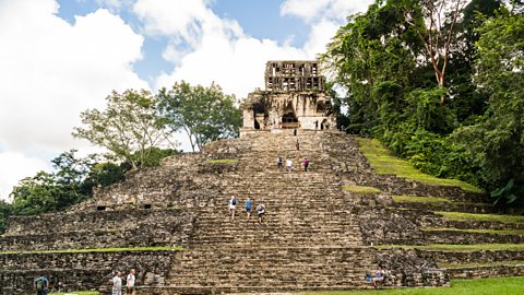 A big stone pyramid with wide steps in the rainforest. People are walking up the steps to a small stone building at the top.
