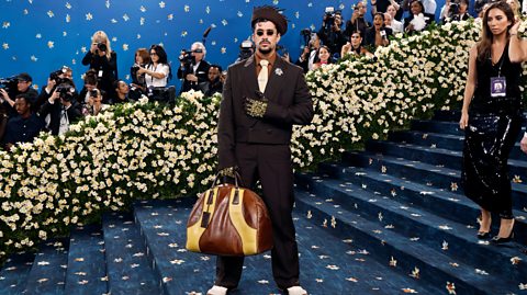 Bad Bunny wears a chocolate brown suit, paired with black sunglasses, a tie, gloves and a traditional pava hat. He holds a large tan and brown leather bag