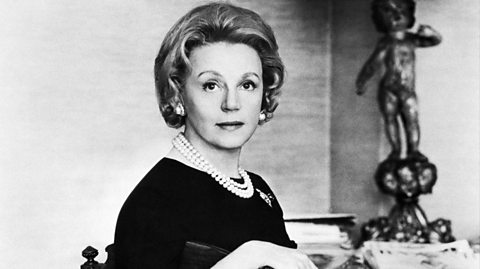 A black and white photo of Eleanor Lambert sitting on a chair looking at the camera. Her arm is resting against the back of the chair and she has short hair, pearl earrings, a pearl necklace, a small broach and wears a long sleeved dark dress