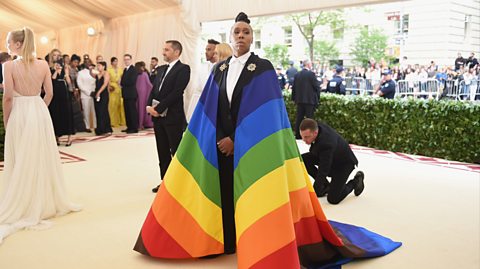 Lena Waithe wearing a Carolina Herrera cape with rainbow colours symbolising the Pride flag. Beneath they wear a black tailored suit with a white shirt, hair in a top knot and stand on a cream carpet