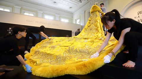 Rihanna's 2015 Met Gala dress by Guo Pei being put on a mannequin by four workers inside a museum. The gown is yellow and gold with embroidered detail on a long train, faux fur lining on the train and a fluffy fur shawl