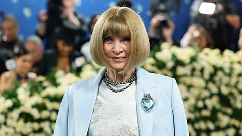 Anna Wintour has bobbed blonde hair with a fringe and wears a cream dress with embroidered detail underneath a light blue suit jacket. She has a chunky silver necklace and light blue broach on her blazer. Blurred behind her is photographers with cameras taking photos