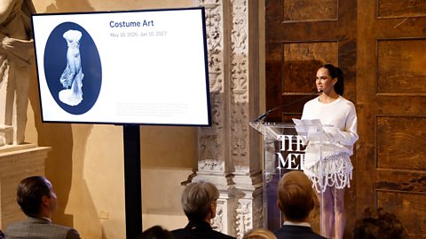 Misty Copeland has dark hair in a ponytail and wears a white long sleeved dress. She stands behind a podium which says 'The Met' and speaks into the mic. Guest watch from her seats in front of her. A large screen next to her reads 'Costume Art' - the theme for the Met's spring exhibition 2026