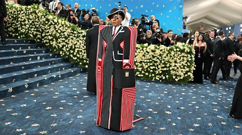  Janelle Monáe attends the 2025 Met Gala celebrating "Superfine: Tailoring Black Style". On the dark blue floral carpet, she wears surrealist outfit with a 1930s overcoat with red and pinstripe patterns to appear as a suit, with a motorized monocle.